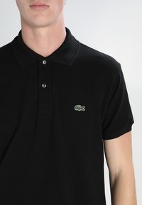 T-shirt polo noir avec un col et une patte de boutonnage à deux boutons. Logo crocodile vert Lacoste brodé sur la poitrine gauche.