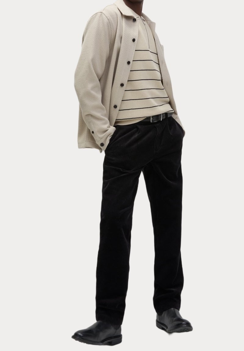 Homme portant une chemise beige boutonnée sur un polo beige rayé, pantalon en velours côtelé noir et chaussures en cuir noires, debout les mains dans les poches.