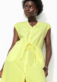 Robe boutonée jaune clair avec des manches courtes, taille cintrée et poches latérales. Tissu doux, longueur genou avec un col en V.