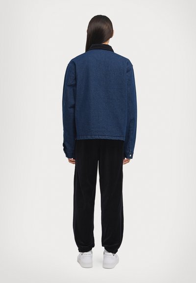 Denim jacket in dark blue with a collar, long sleeves, and button closures, paired with black fleece pants and white sneakers. Back view.