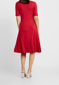 Punainen lyhythihainen mekko, jossa on tiukka yläosa ja levenevä hameosa. Sujuva rakenne, polvipituinen, yhdistetty vaaleisiin tukkijalkapohjaisiin sandaaleihin.