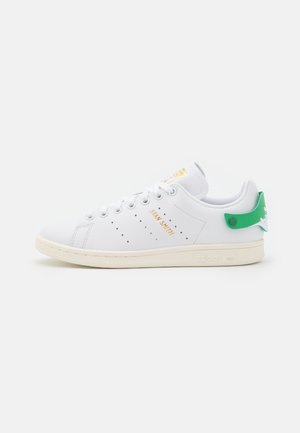 Zapatilla blanca Adidas Stan Smith con lengüeta verde en el talón, logotipo dorado y suela blanco roto, vista de lado sobre fondo blanco.