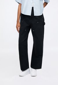 Μαύρο παντελόνι φαρδύ καμπάνα από τζιν ύφασμα, με καμπυλωτές τσέπες διακοσμημένες με μεταλλικά στοιχεία, σε συνδυασμό με λευκά αθλητικά παπούτσια σε ουδέτερο φόντο.