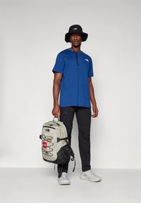 Mėlyna trumpomis rankovėmis marškinėliai, juodos kelnės su kišenėmis, pilka kuprinė su raudonomis detalėmis; balti sportiniai bateliai; juoda kibiro formos kepurė; stovima poza.