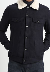 Granatowa kurtka z kołnierzem z owczej wełny, zapinana na guziki, z dwiema kieszeniami na piersi i ściągaczami na rękawach. Teksturowany materiał z pionowymi szwami.