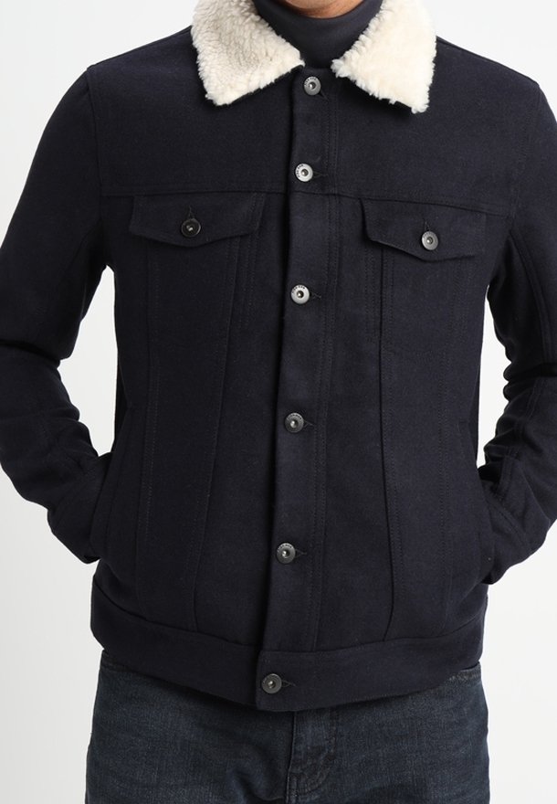 Granatowa kurtka z kołnierzem z owczej wełny, zapinana na guziki, z dwiema kieszeniami na piersi i ściągaczami na rękawach. Teksturowany materiał z pionowymi szwami.