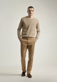 Joven de pie con las manos en los bolsillos, vistiendo un suéter beige de cuello en V, pantalones caqui y zapatos marrones de deslizarse en un fondo liso.