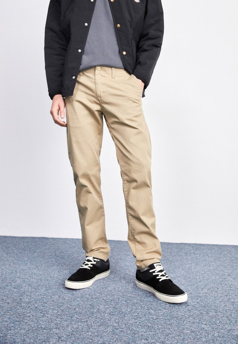 Homme portant un pantalon beige, des baskets noires avec des lacets blancs, une chemise grise et une veste noire debout sur un tapis bleu.