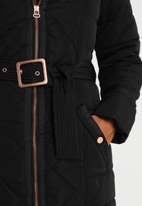 Manteau matelassé noir avec une fermeture éclair cuivre, une boucle de ceinture carrée assortie, et une main glissée dans une poche latérale avec un bouton cuivre.