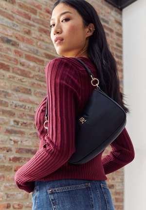 Mujer con un suéter rojo de canalé y jeans azules sosteniendo un bolso negro con herrajes dorados sobre el hombro, frente a una pared de ladrillos.