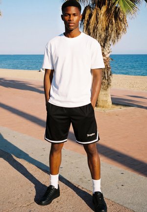 Jeune homme portant un t-shirt blanc, un short noir et des baskets noires, debout sur un chemin en bord de mer avec un palmier et l'océan en arrière-plan.
