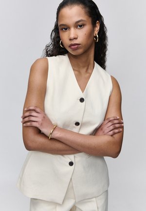 Femme bras croisés, portant un gilet beige sans manches et un pantalon assorti, des boucles d'oreilles créoles dorées et un bracelet en or, posant sur fond gris.
