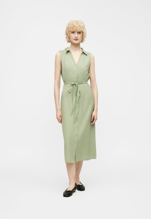 Γυναίκα με μπούκλες ξανθά μαλλιά, που φοράει αμάνικο midi φόρεμα ανοιχτού πράσινου χρώματος με κουμπιά, ζώνη στη μέση και μαύρα μπαλαρίνα παπούτσια, στέκεται σε λευκό φόντο.