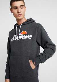 Mikina s kapucí v barvě uhlově šedé má přední kapsu, bílé šňůrky a výrazné logo ellesse v bílé a oranžové barvě.