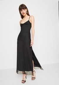 Black slip dress with thin straps, thigh-high slits, and a smooth, glossy fabric. Worn with strappy black heels and statement earrings.