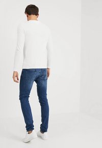 Vit långärmad skjorta kombinerad med mörkblå slim-fit jeans och vita sneakers, sedd bakifrån mot en enfärgad ljus bakgrund.