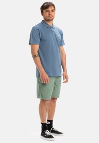 Ljusblå polo med korta ärmar och krage, tillsammans med gröna shorts och svarta sneakers. Enkelt mönster och bomullstextur.