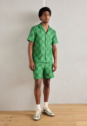 Chemise à manches courtes à motifs verts et shorts assortis, fabriqués en tissu léger à motifs carrés. Portés avec des baskets vertes et grises.