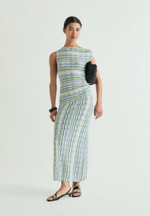 Mujer con vestido midi sin mangas con estampado de zigzag multicolor, sandalias negras, brazalete negro y sujetando un bolso negro bajo el brazo.