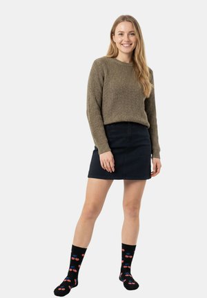 Ung kvinde smilende, iført brun strikket sweater, sort denim mininederdel og sorte sokker med rødt kirsebærmønster, stående på hvid baggrund.