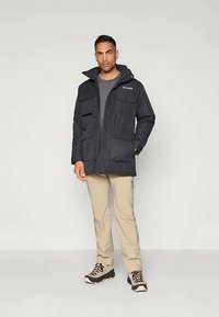 Ναυτικό εξωτερικό μπουφάν με πολλαπλές μπροστινές τσέπες, σχέδιο με φερμουάρ και ρυθμιζόμενη κουκούλα. Συνδυασμένο με ανοιχτού χρώματος καφέ παντελόνι και μποτάκια πεζοπορίας.