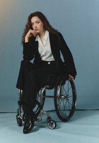Blazer de terciopelo negro sobre una camisa blanca, combinado con pantalones negros y zapatos negros brillantes, sentado en una silla de ruedas moderna contra un fondo azul.