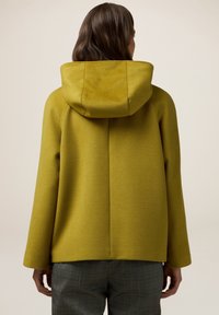 Cappotto a cappuccio giallo-verde con una finitura testurizzata, caratterizzato da un taglio dritto e maniche larghe, vista da dietro.
