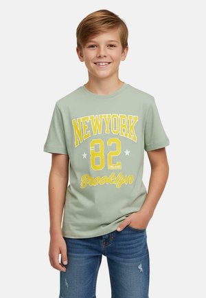 Groen katoenen T-shirt met "NEW YORK 82" en "Brooklyn" in gele letters, korte mouwen, ronde halslijn, gedragen met blauwe spijkerbroek.