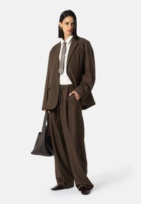 Bruine oversized blazer en bijpassende broek met een taille met trekkoord, gecombineerd met een wit overhemd en geruite stropdas. Inclusief zwarte handtas en schoenen.