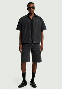 Camisa de manga curta em denim preto com calções combinando, apresentando um bolso e botões de metal. Usada com meias e sapatos pretos, e óculos de sol.