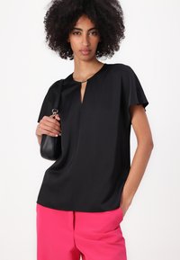 Zwart kortemouwtop met een keyhole halslijn, gladde textuur, gedragen met roze broek, aangevuld met een kleine zwarte handtas.