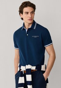 Giovane uomo che indossa una polo blu navy con bordi bianchi e logo "Façonnable 1950", con maglione a righe annodato in vita.