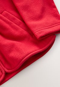 Gros plan d'une manche pliée d'une veste polaire rouge montrant un tissu doux texturé et un ourlet cousu sur fond blanc.