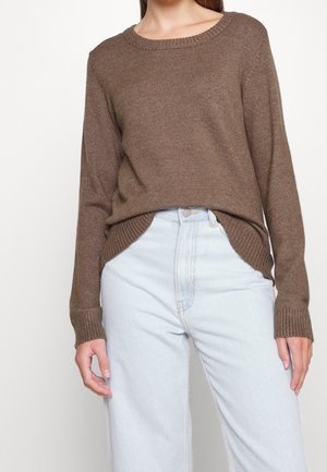 Femme portant un pull en maille marron légèrement rentré dans un jean denim bleu clair taille haute, debout les mains détendues le long du corps.