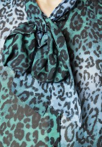 Blouse en satin avec imprimé léopard vert et bleu, détail d'écharpe nouée au niveau du col, avec une texture lisse et légère.