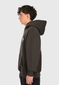 Junger Mann mit lockigem Haar, der einen dunklen, übergroßen Hoodie mit einer Fronttasche trägt, steht im Profil vor einem schlichten hellen Hintergrund.