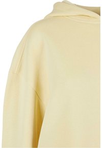 Jasnożółta bawełniana bluza z kapturem, ukazująca detale ramion i górnej części klatki piersiowej, z widocznym szwem i złożonym kapturem.
