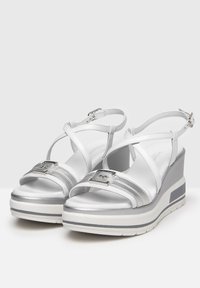 White wedge sandals with a shiny finish, featuring crisscross straps and silver accent bands. Buckle closure, textured sole for grip.