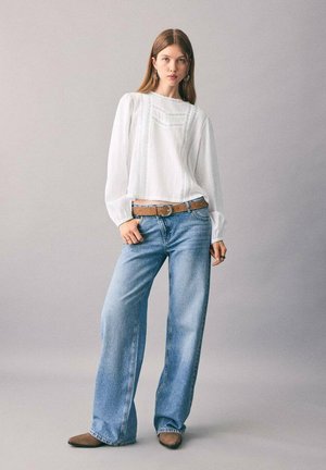 Blusa blanca de manga larga con tejido texturizado y un pequeño bolsillo, combinada con jeans de pierna ancha de color azul claro y cinturón marrón, llevada con zapatos marrones.