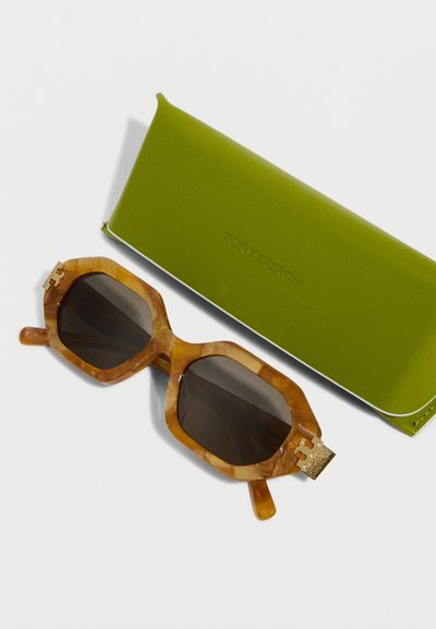 Lunettes de soleil hexagonales en écaille de tortue avec des verres foncés, posées à côté d'un étui à lunettes Tory Burch vert olive sur un fond blanc.