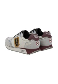 Un paio di sneaker Aeronautica Militare bianche e grigie con dettagli bordeaux, lacci neri, toppe militari e il tondo italiano sul tallone.