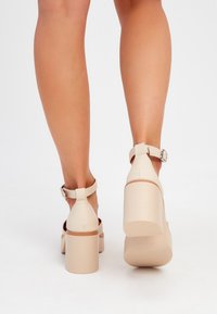 Beige högklackade sandaler med en kraftig klack, justerbart ankelsnöre, slät yta och öppen baksidesdesign.