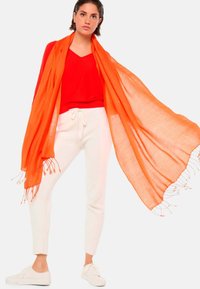 Écharpe orange drapée sur un haut rouge à col en V, associée à un pantalon blanc avec cordon de serrage et des baskets blanches. L'écharpe présente des franges.