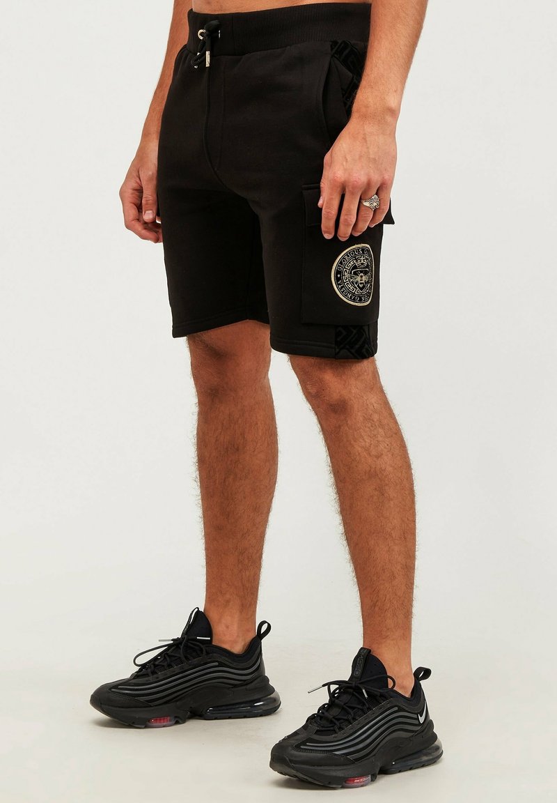 Homme portant un short cargo noir avec un patch circulaire et des chaussures de sport noires, debout sur un fond clair uni.