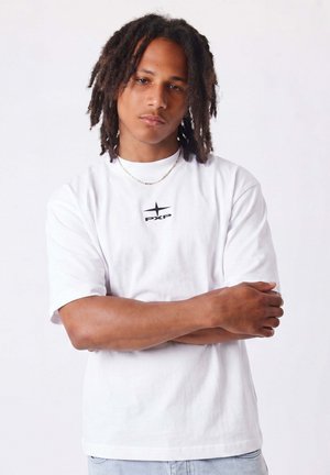 Giovane uomo con pelle medio-scura e dreadlocks, in piedi con le braccia incrociate, indossa una maglietta bianca con il logo PXP e una sottile collana a catena.