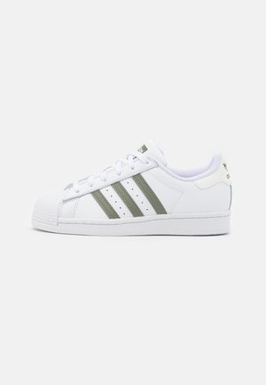 Hvid Adidas-sneaker med olivengrønne striber og skalkappe, set fra ydersiden på en ensfarvet lys baggrund.