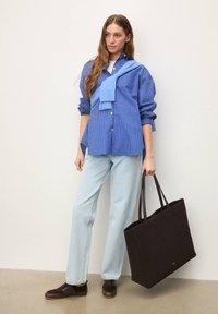 Chemise à rayures bleues et blanches à boutons, pull bleu clair posé sur l'épaule, jean bleu clair, chaussures marron foncé, sac fourre-tout noir.