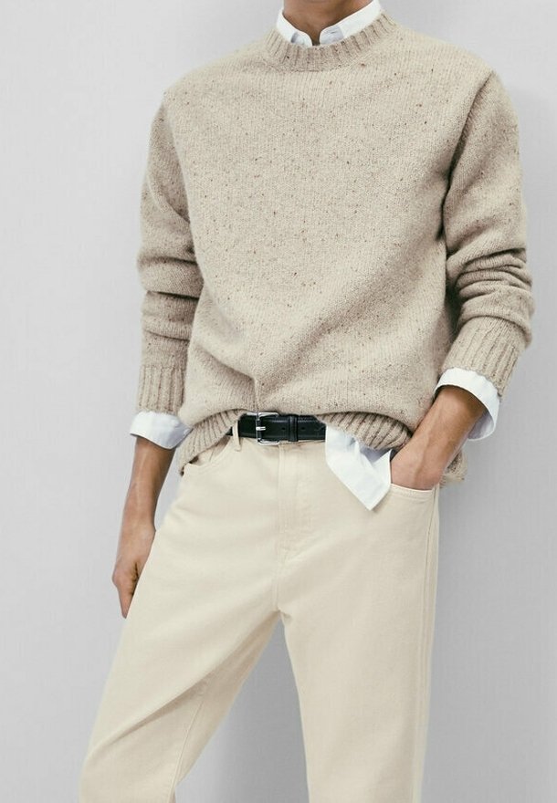 Homme portant un pull en maille beige sur une chemise blanche rentrée dans un pantalon beige clair avec ceinture noire, une main dans la poche.