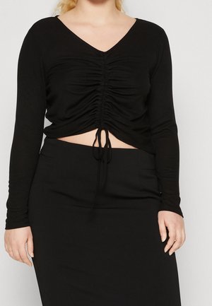Vrouw die een zwart gerimpeld cropped top met lange mouwen en verstelbare trekkoorden draagt en een zwarte rok, staand voor een effen achtergrond.