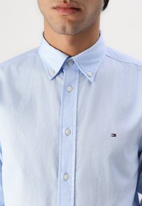 Camisa abotonada de color azul claro con cuello, hecha de una tela con un acabado ligeramente texturizado. Presenta un pequeño logo en el lado izquierdo del pecho.
